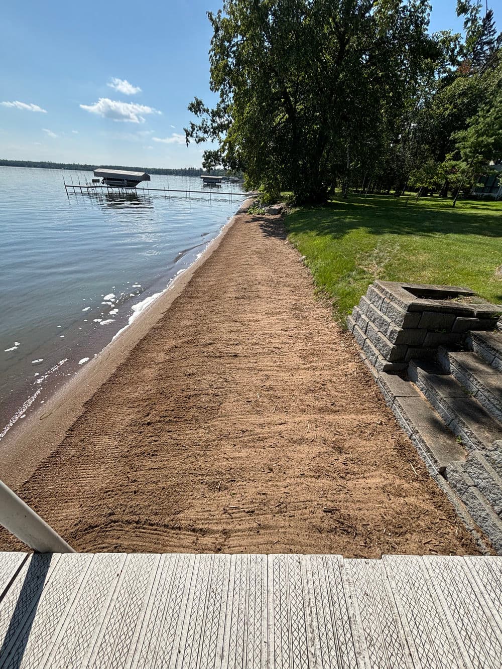 Tranquil lakeside view with sandy shore, docked boats, and green lawn under clear blue sky.