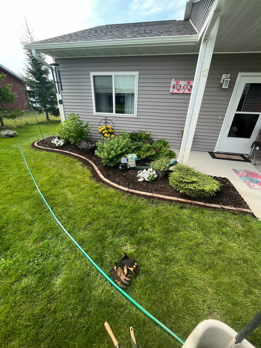 Lush garden bed with colorful flowers and welcome sign near a house exterior.