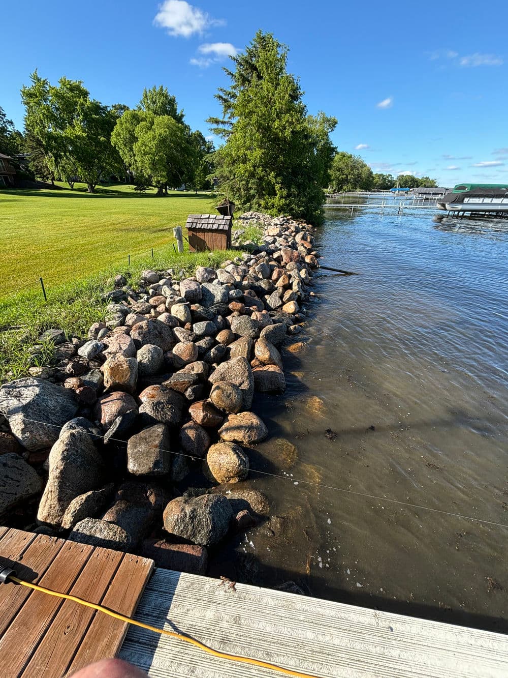 Rocky shoreline along a calm lake with green trees and a wooden dock visible.